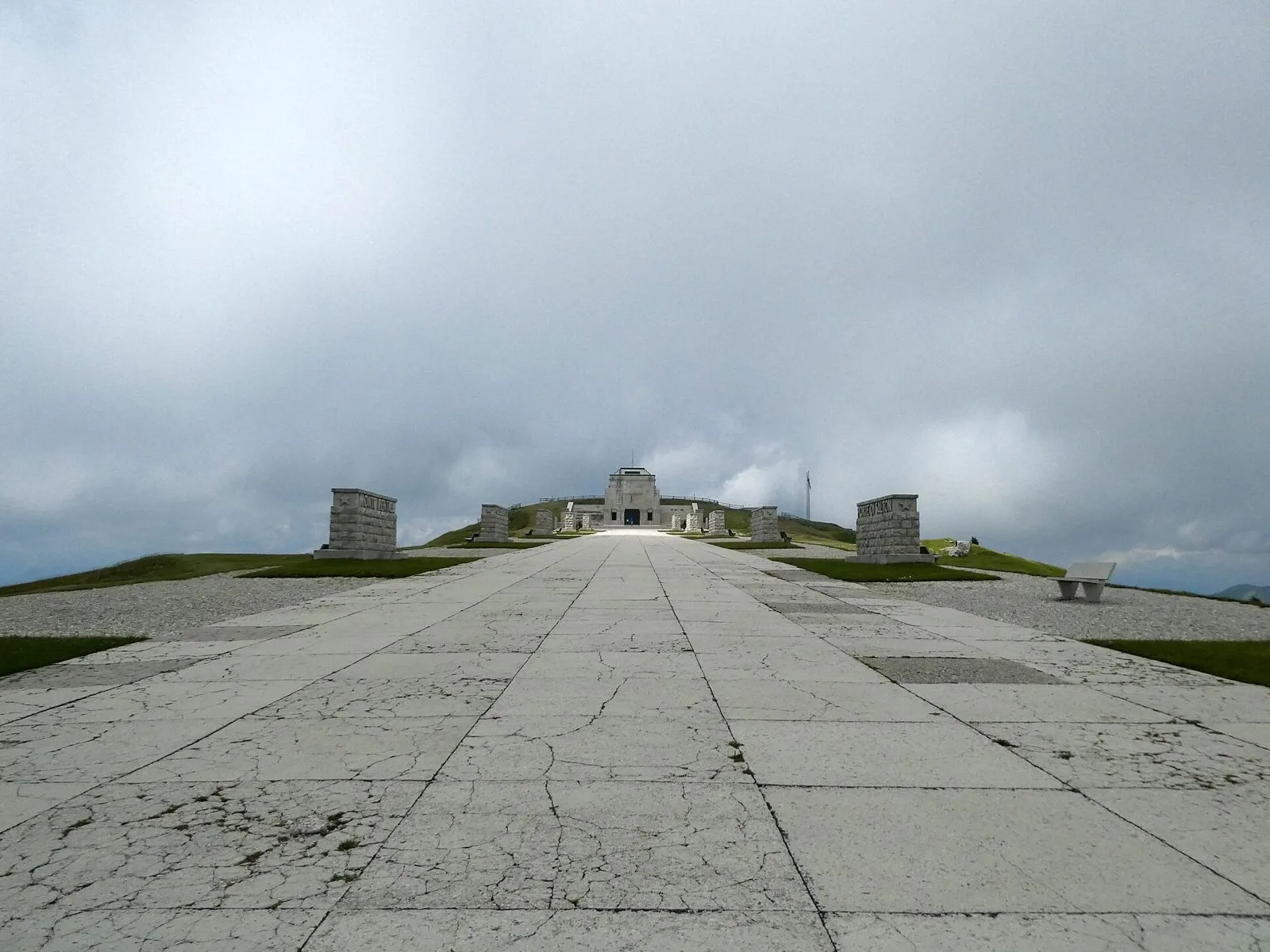 La Via Eroica del Sacrario Militare di Cima Grappa, viale lastricato fiancheggiato da cippi in pietra che conduce all'edificio sommitale sotto un cielo nuvoloso